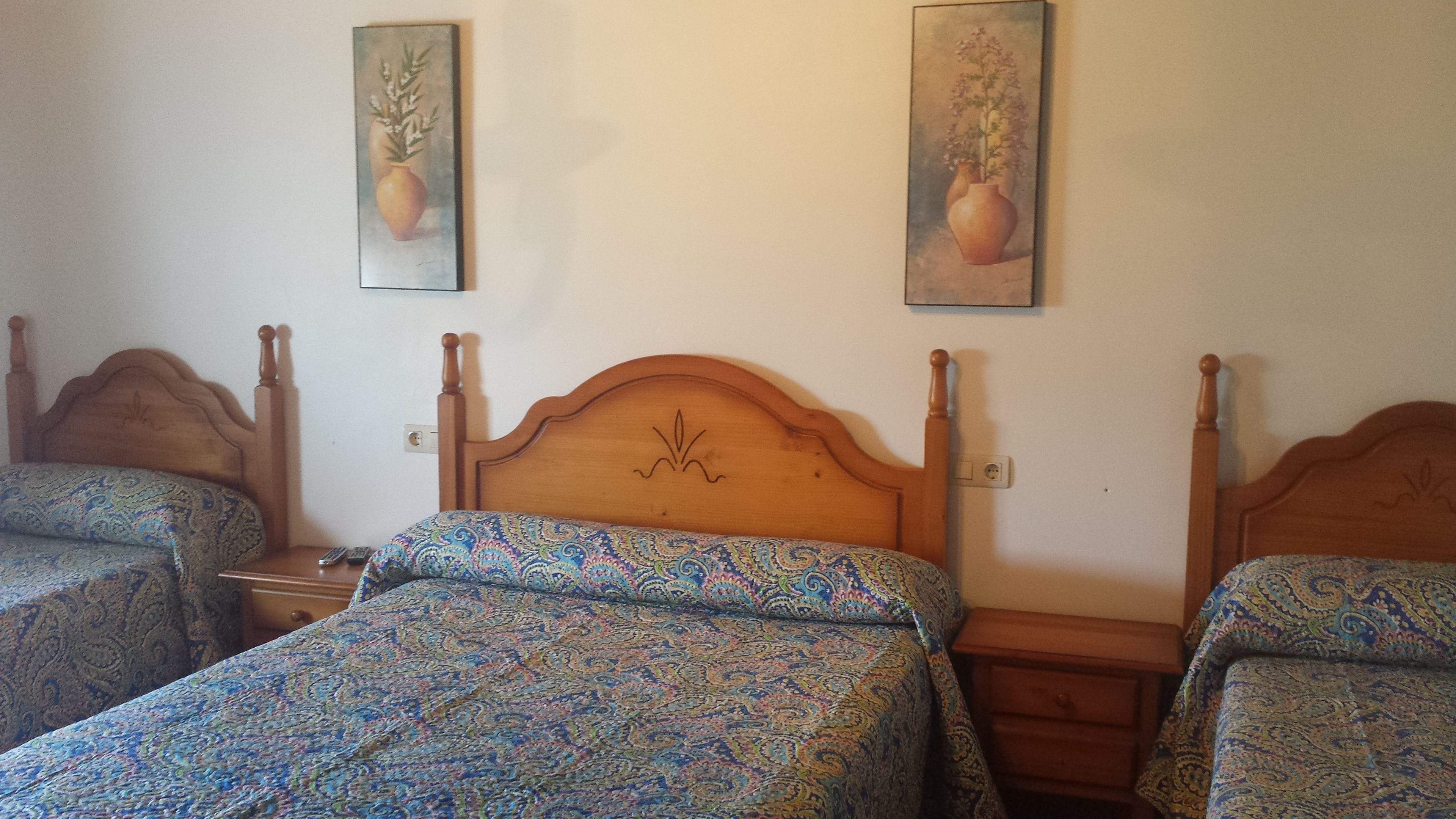 A room with two beds, each with a patterned bedspread, wooden headboards, and a nightstand with a lamp. There are two paintings on the wall above the beds.