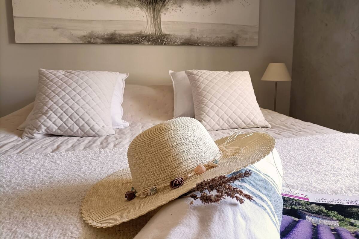 A bed with white bedding, a white quilted pillow, a straw hat, and a painting of a tree on the wall.