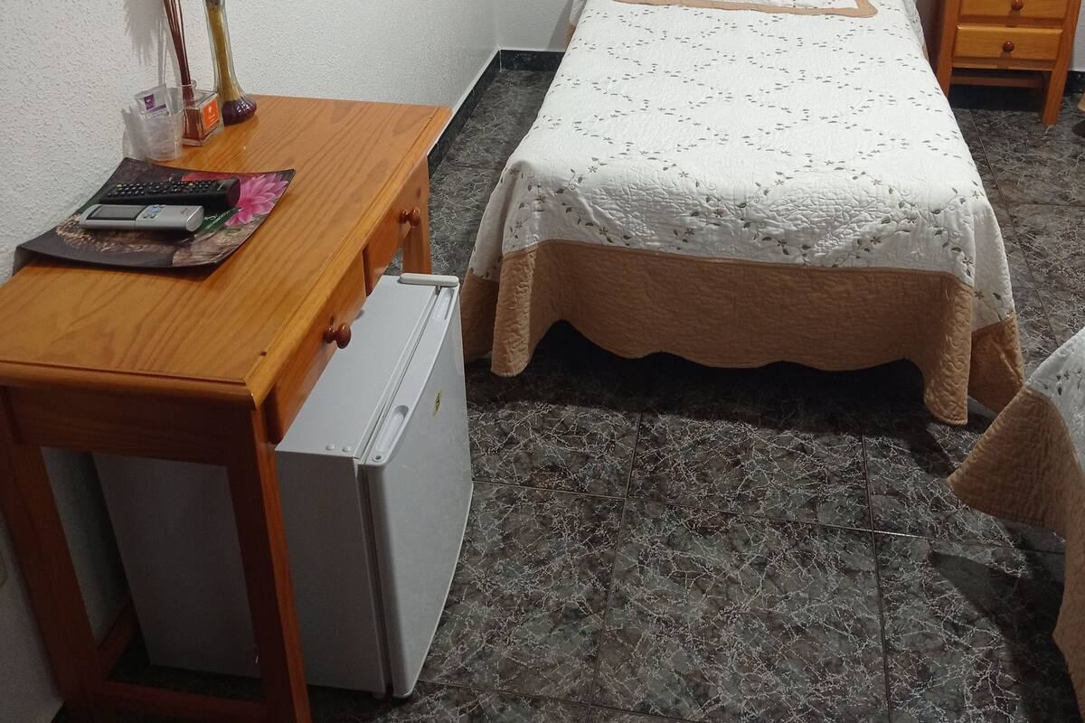 A bedroom with a wooden bed, a small desk with a refrigerator, a nightstand with a lamp, and a wall with framed pictures.