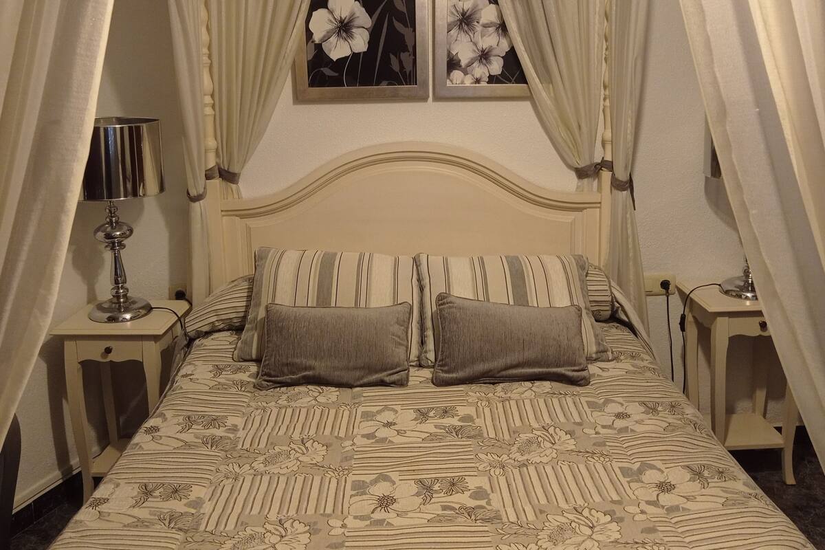 A bedroom with a bed, two pillows, a nightstand with a lamp, and a painting on the wall.