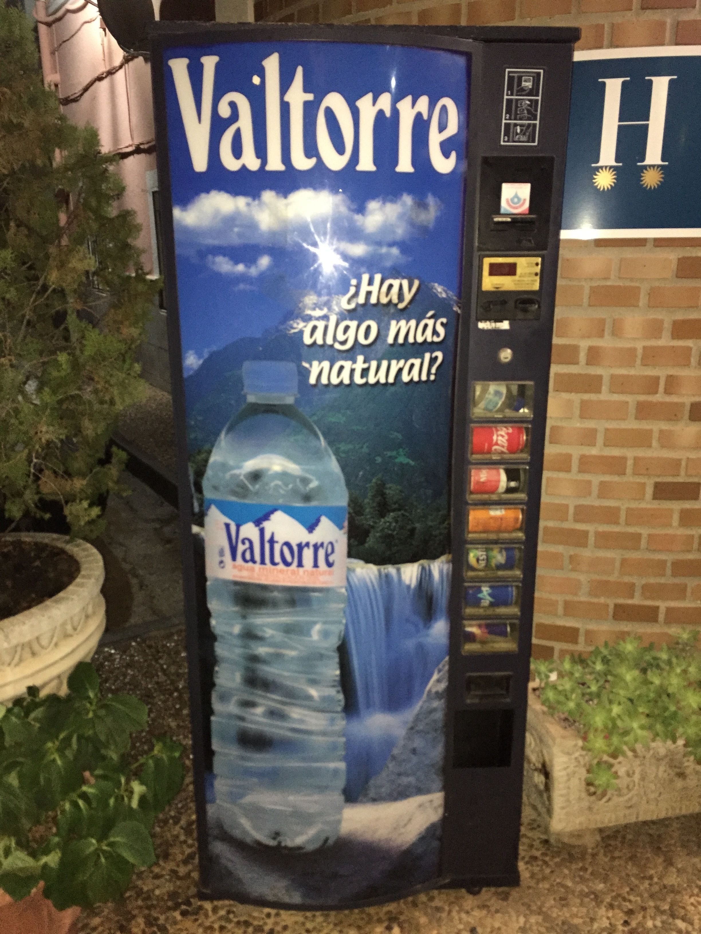 Un distributeur d’eau Valtorre, orné d’une publicité représentant un paysage naturel, est installé à l’extérieur, près d’un mur de briques.