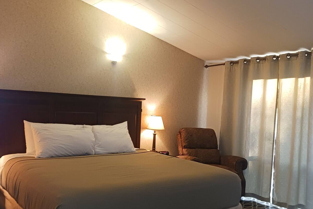A hotel room with a large bed, a nightstand, a lamp, a chair, and a window with curtains.