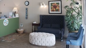 Un espacio de sala acogedor con un sofá capitonado, un reposapiés redondo, un sillón azul, un mostrador de recepción verde, un reloj, una obra de arte floral enmarco y una planta en maceta.