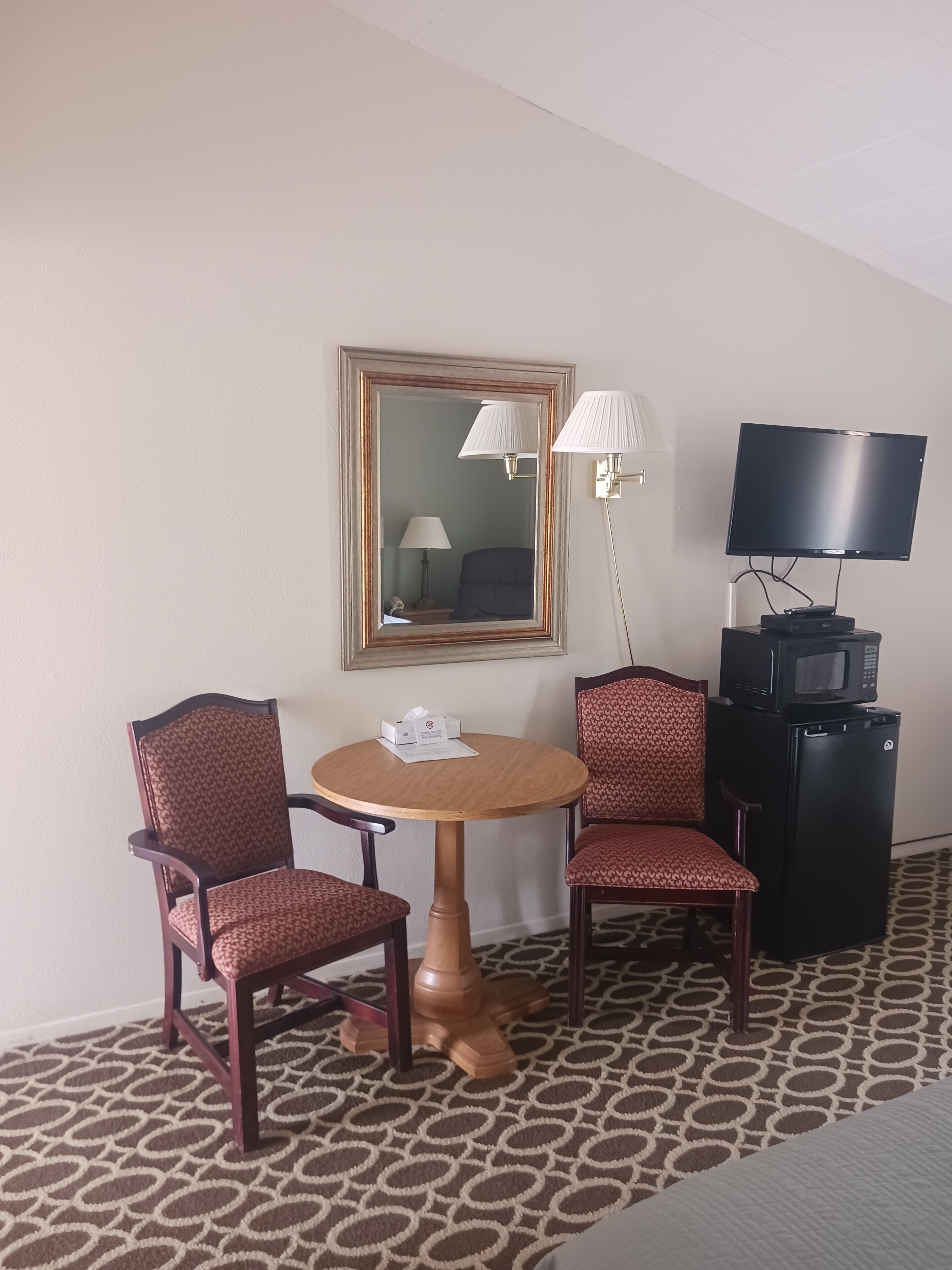 A hotel room with a round wooden table, two patterned chairs, a television, a microwave, and a lamp.