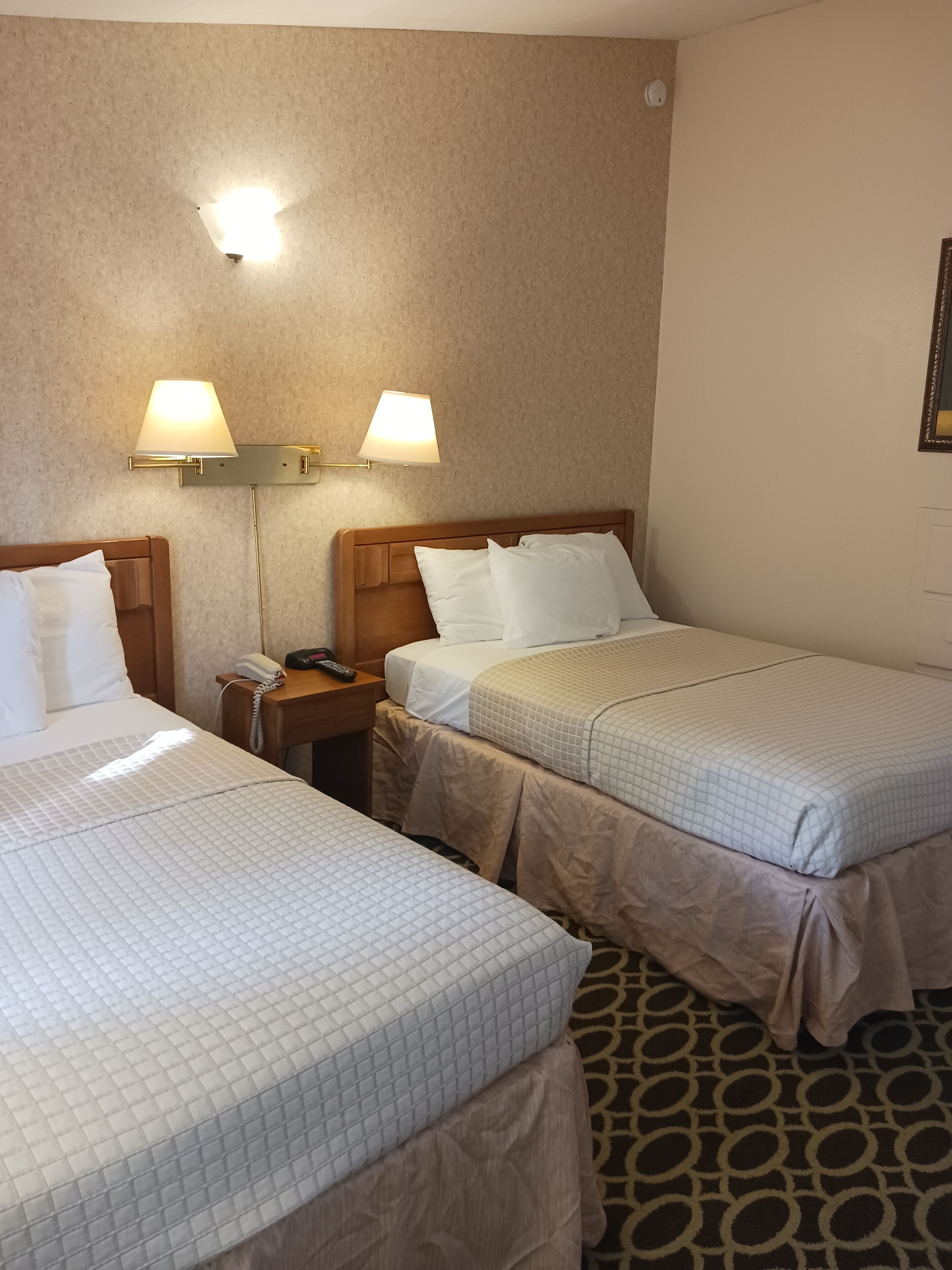 A hotel room with two beds, each with a white quilted bedspread, a wooden headboard, and a bedside table with a lamp.