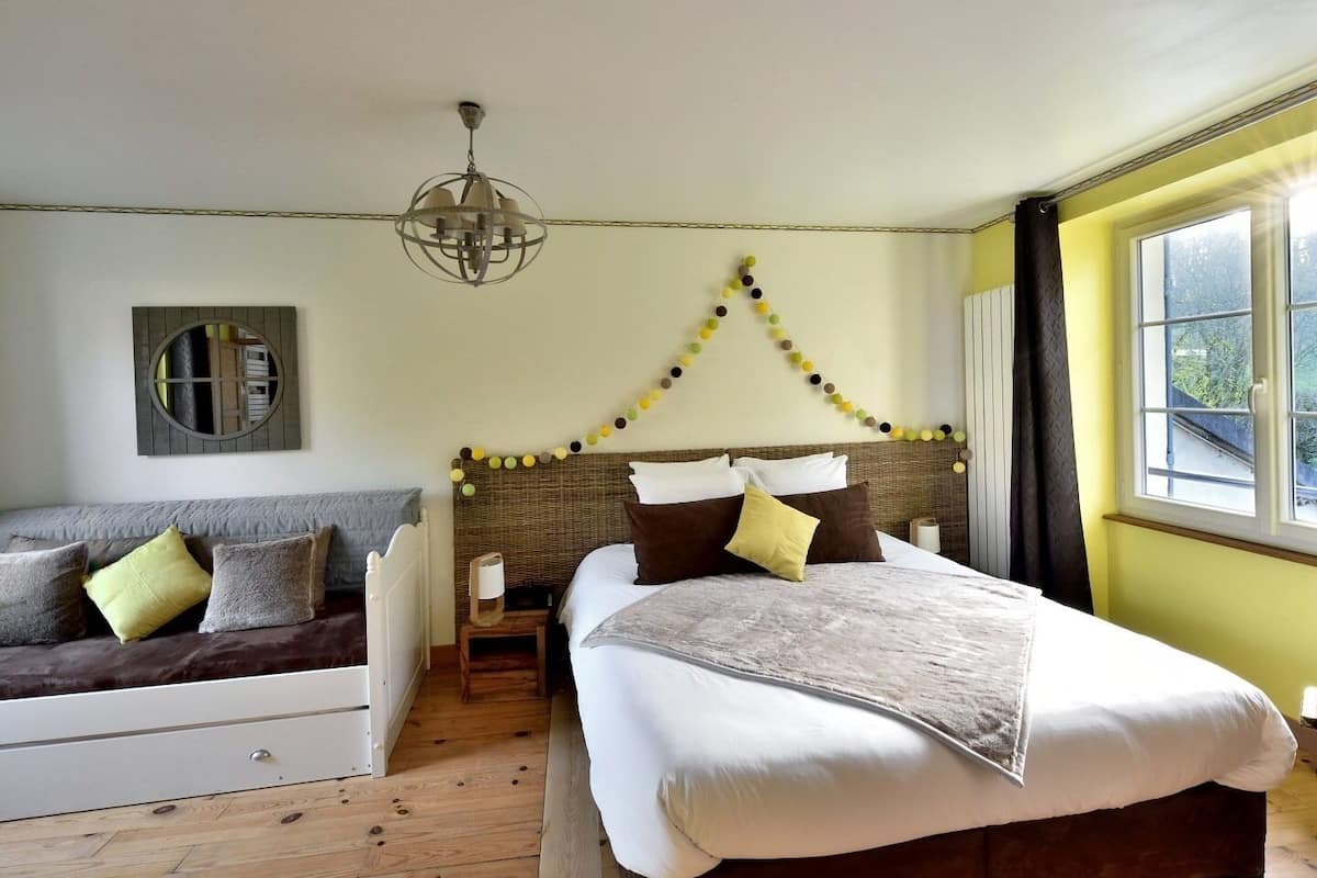 A bedroom with a bed, a sofa, a wooden floor, a window, and a wall-mounted mirror.