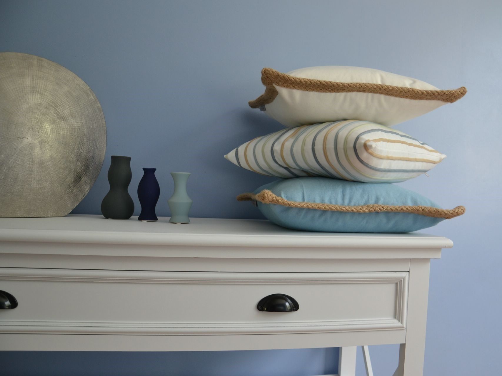 Une commode blanche avec un tiroir, un grand miroir rond et trois vases de différentes hauteurs et couleurs.