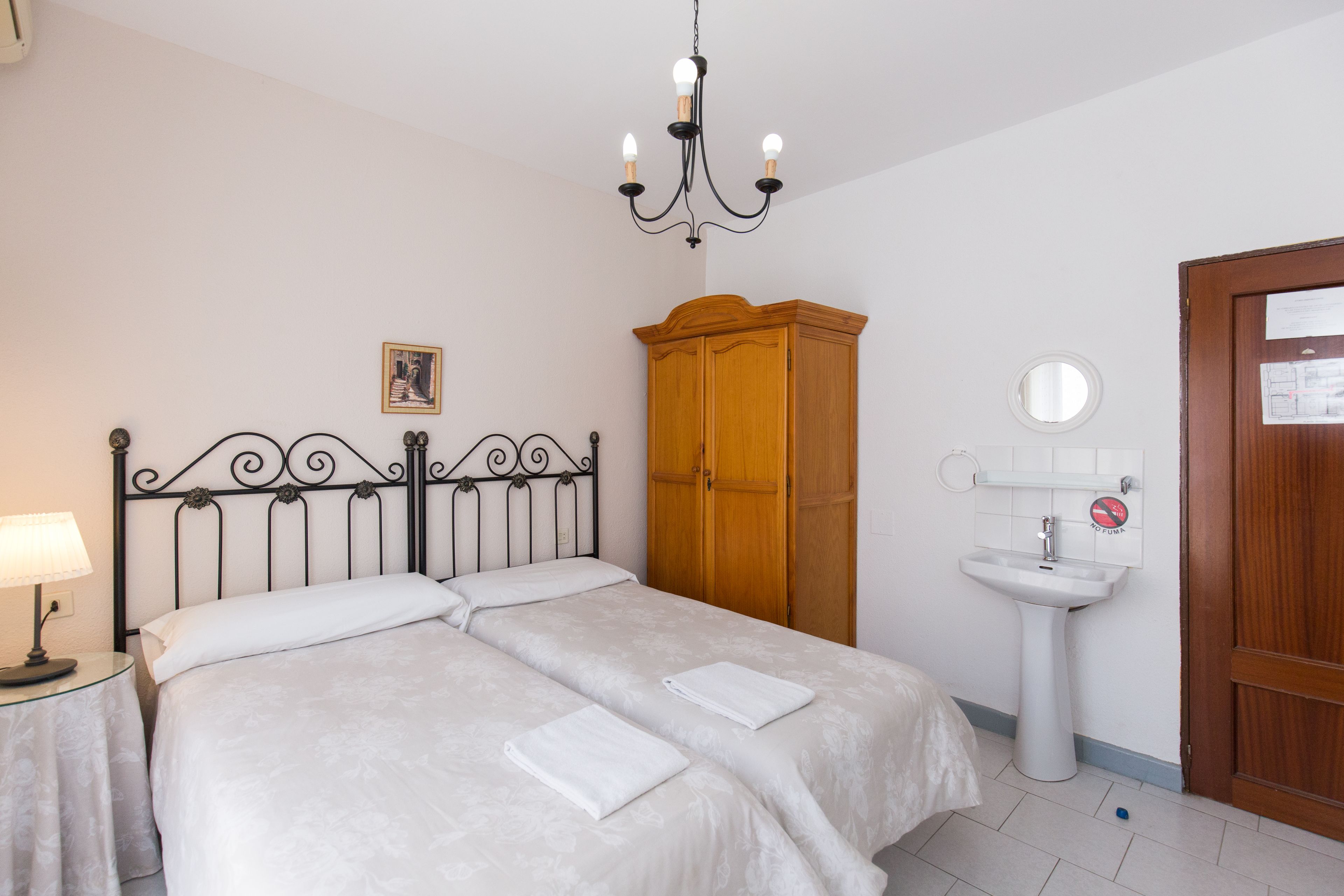 Un llit doble amb dos almoines, una taula de nit amb una làmpada, un armari de fusta, un lavabo blanc i un mirall rodó a la paret.
