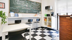 Habitación de hotel con piso a cuadros, un escritorio con dos sillas, una pizarra con información cultural, una recepción y una pequeña kitchenette.