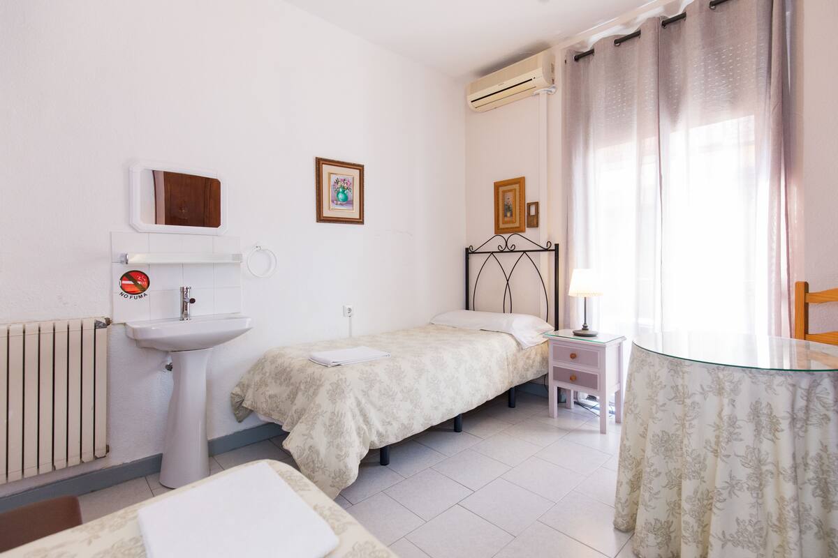 A bedroom with a bed, a bedside table with a lamp, a window with curtains, a radiator, a sink, and framed pictures on the wall.