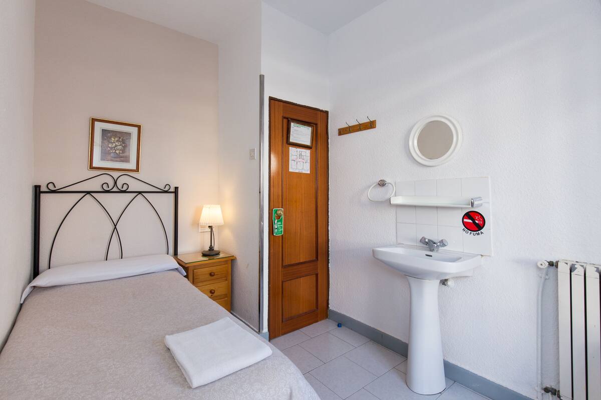 A hotel room with a bed, a bedside table with a lamp, a wooden door, a bathroom with a sink and mirror, and a radiator.