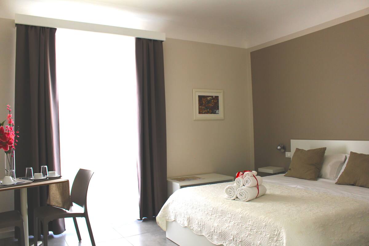 Ein Hotelzimmer mit Bett, Schreibtisch mit Stuhl, einem kleinen Tisch und einem Fenster mit Vorhängen.