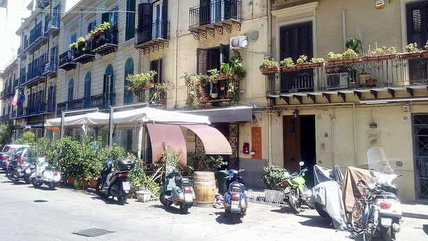 A row of multi-story buildings with balconies, some with plants. There are parked scooters and a few cars. - Triscele Glamour Rooms (Palermo)