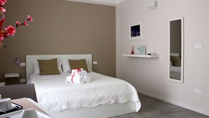 A neatly arranged hotel room with a large bed, a bedside table with a lamp, a small shelf with toiletries, and a mirror on the wall. - Triscele Glamour Rooms (Palermo)