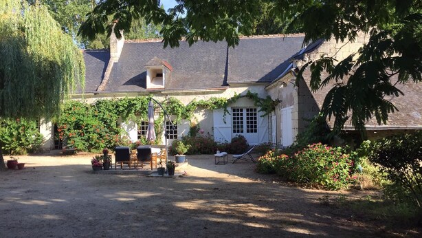 A rustic house with a gravel driveway, a wooden deck with chairs, and a small garden area with potted plants and flowers. - Le Moulin Bertrand (Benais)