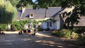 A rustic house with a gravel driveway, a wooden deck with chairs, and a small garden area with potted plants and flowers. - Le Moulin Bertrand (Benais)
