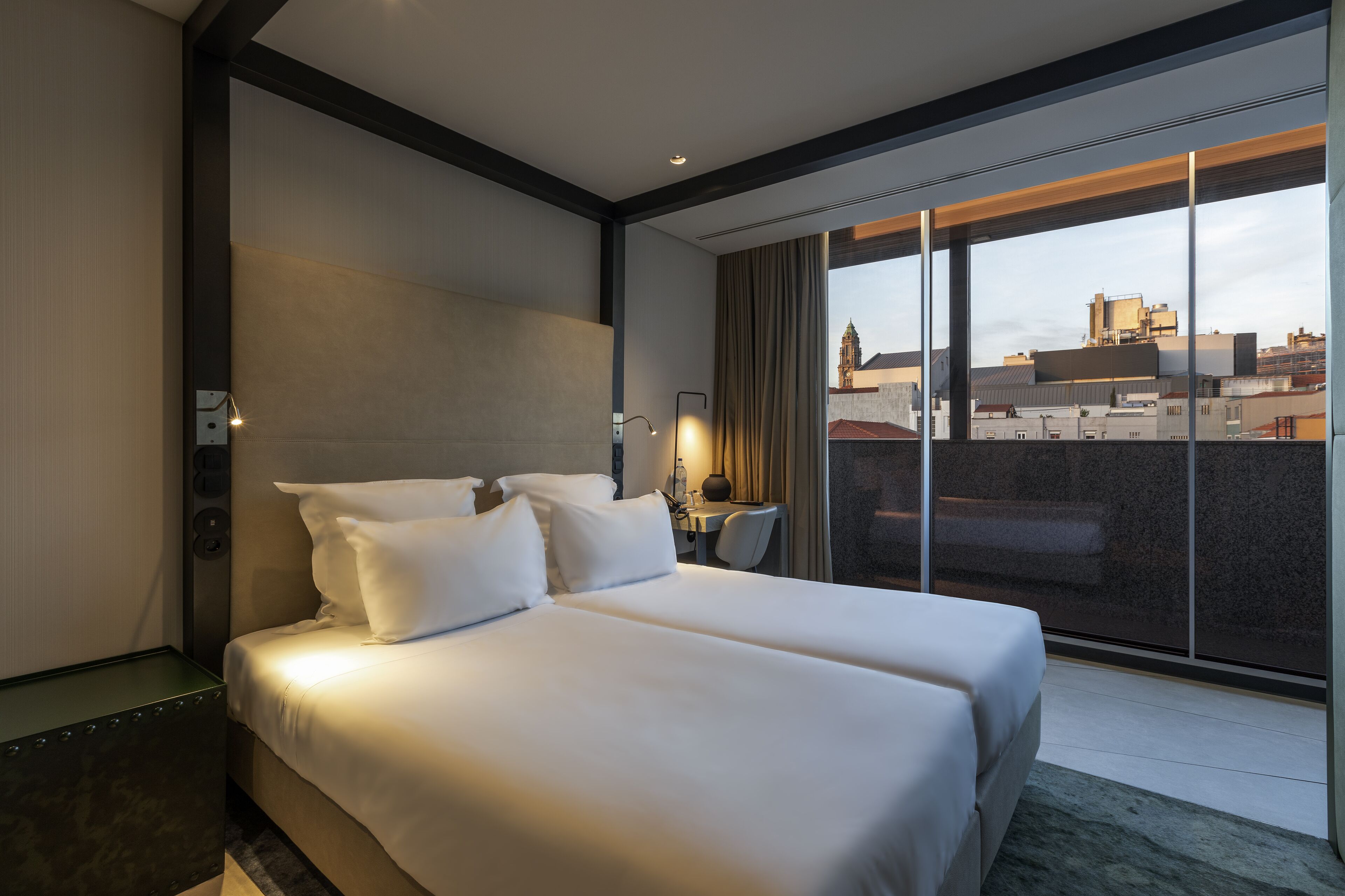 Quarto de hotel com uma cama grande, uma escrivaninha com cadeira e vista da cidade através de amplas janelas.