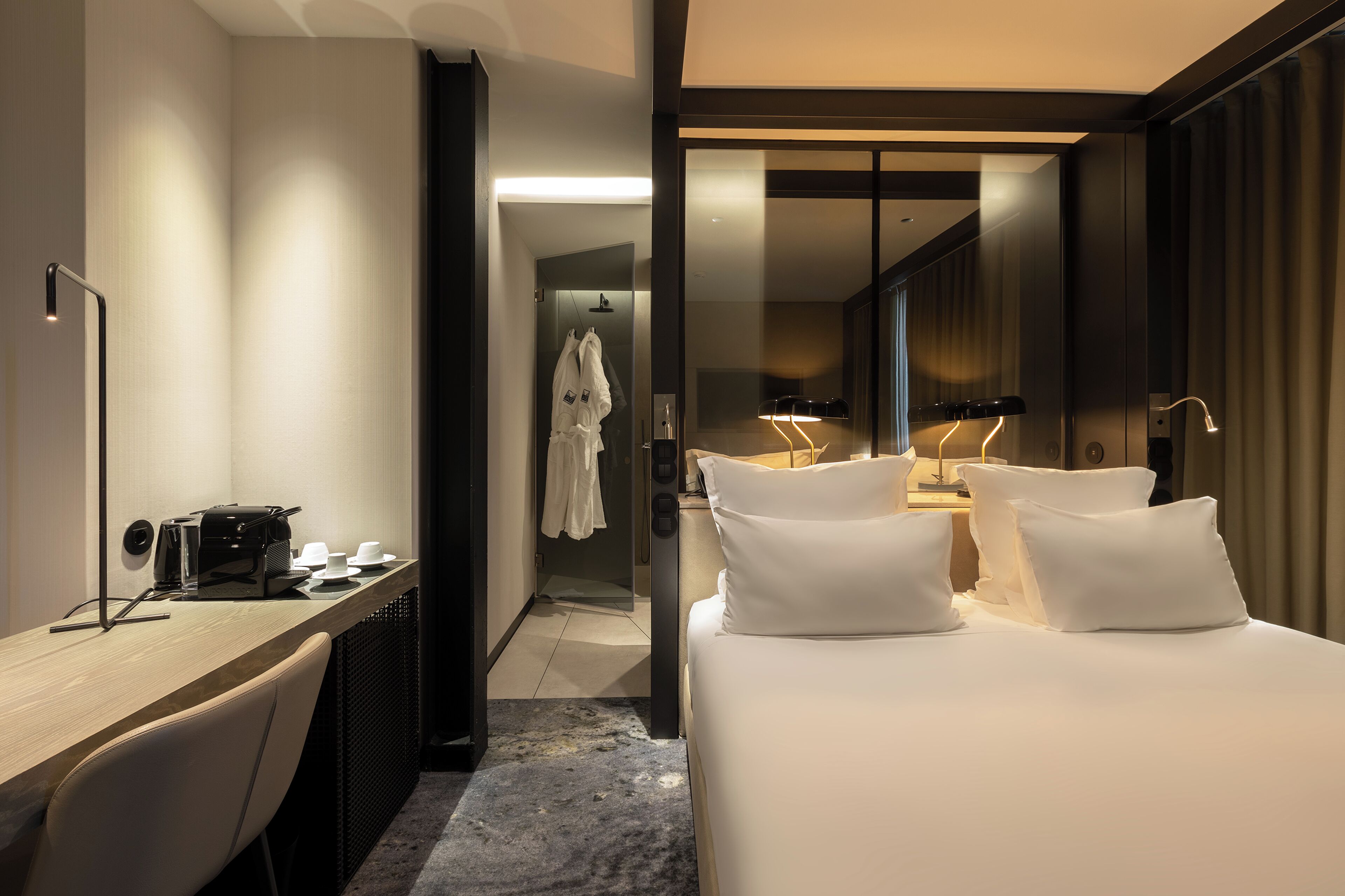 Une chambre d’hôtel moderne équipée d’un bureau, d’un lit avec du linge de maison blanc, d’un espace douche et d’un miroir.