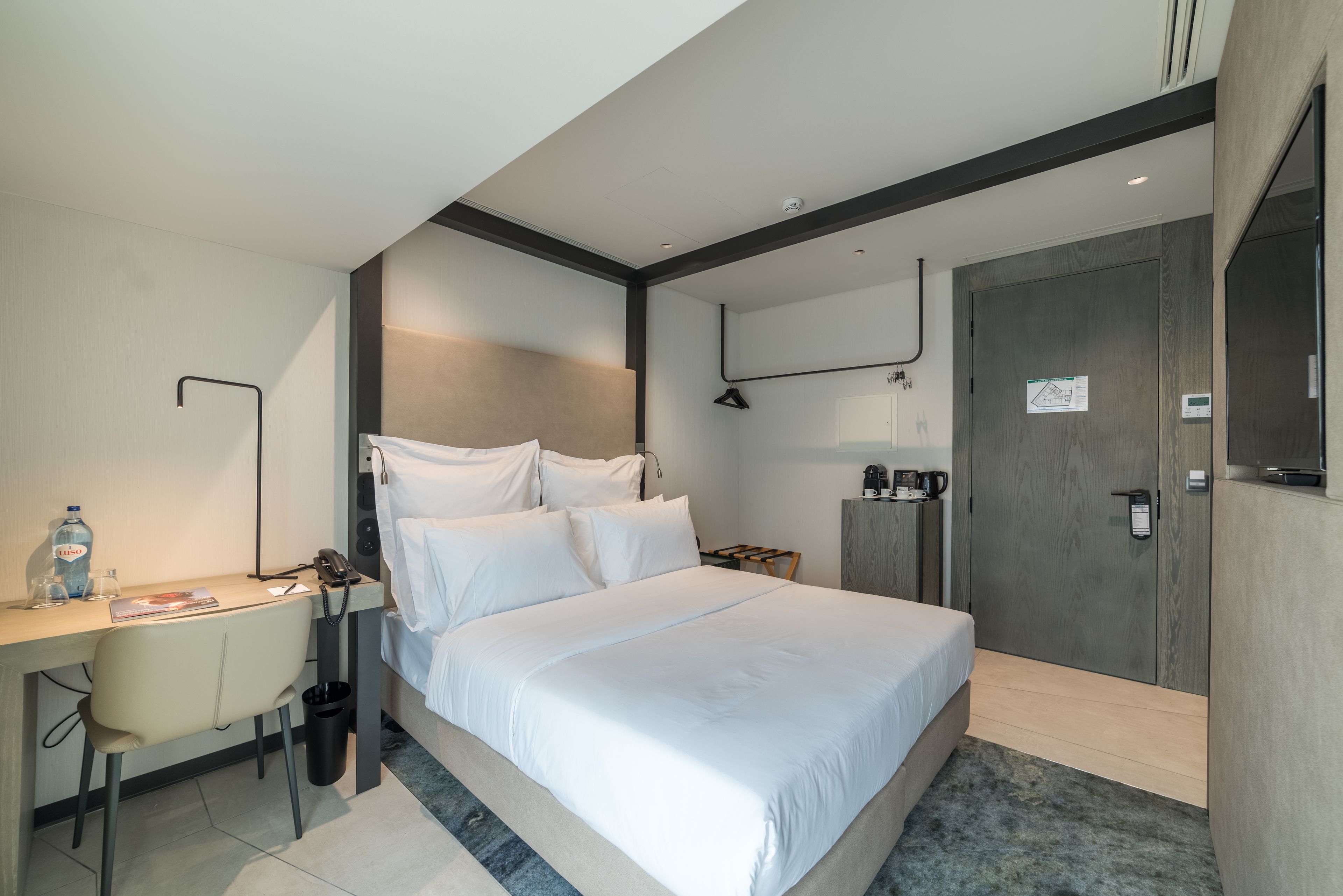 A modern hotel room with a large bed, a desk with a chair, a small table, and a wardrobe.