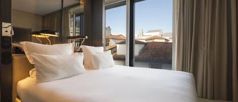 A hotel room with a large bed, a desk, and a view of buildings through a large window.