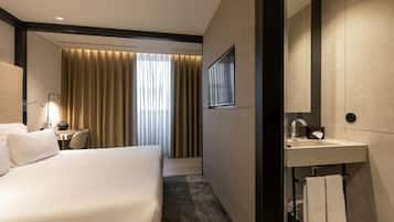 A modern hotel room with a large bed, a desk, a desk lamp, a bathroom with a sink and mirror, and a view of the outside through curtains.