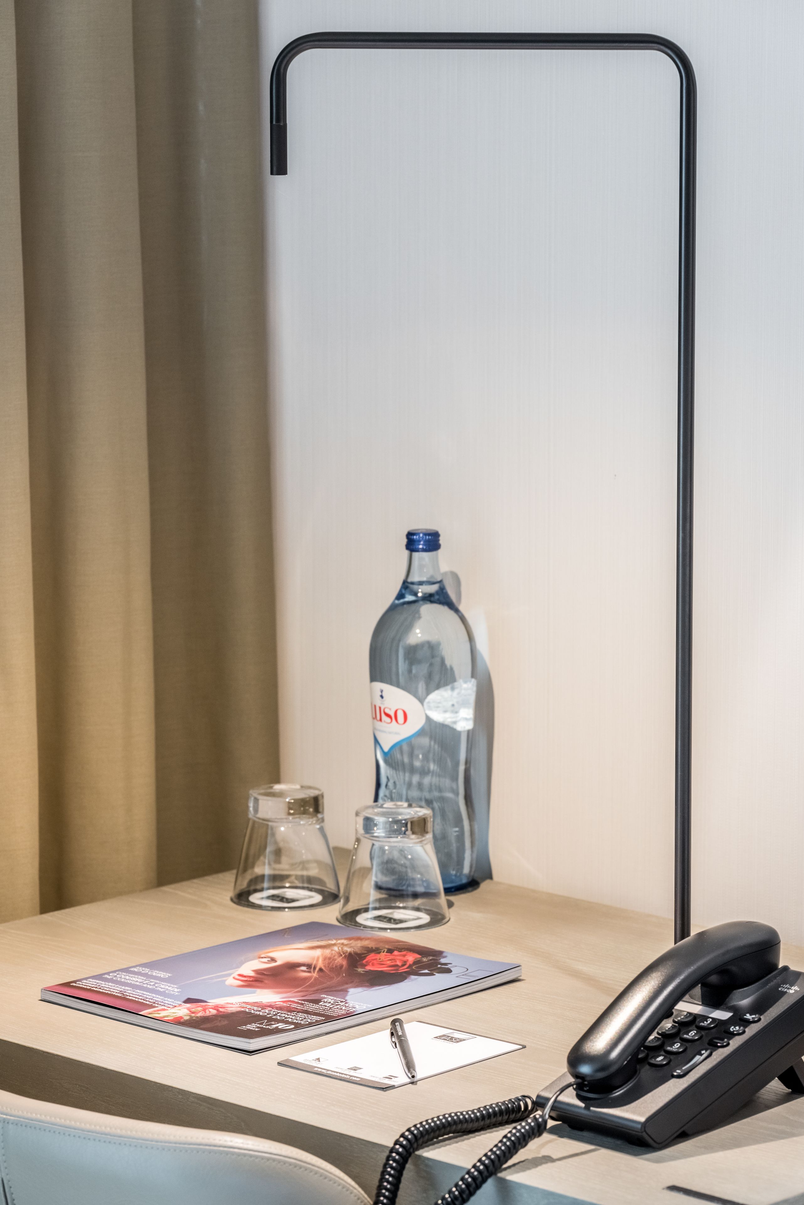 A hotel room with a desk, a telephone, a bottle of water, and a glass.