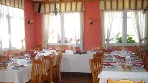 A dining area with multiple tables set for a meal, wooden chairs, and large windows with curtains. - Pension Restaurante Vazquez (Vimianzo)