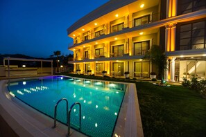 Un hotel de varios pisos con piscina, mobiliario exterior e iluminación destacada en la fachada.
