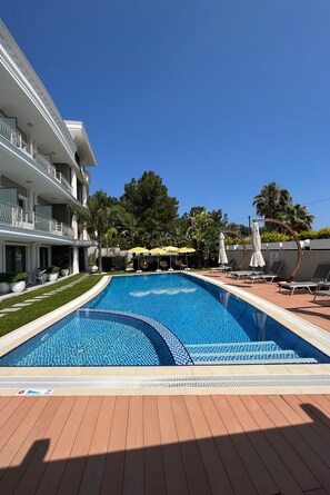 Una piscina con sillas de descanso y sombrillas, rodeada por un edificio con balcones y un césped bien cuidado.