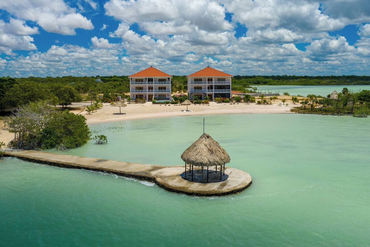 Un complexe hôtelier comprenant deux grands bâtiments, une plage et une structure à toit de chaume située dans l’eau.