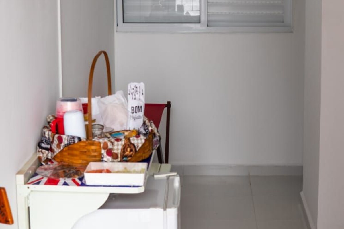 Um quarto pequeno e estreito, com chão de azulejos brancos, uma janela com persianas, um ar condicionado fixado na parede, uma televisão e uma mesa com diversos objetos.