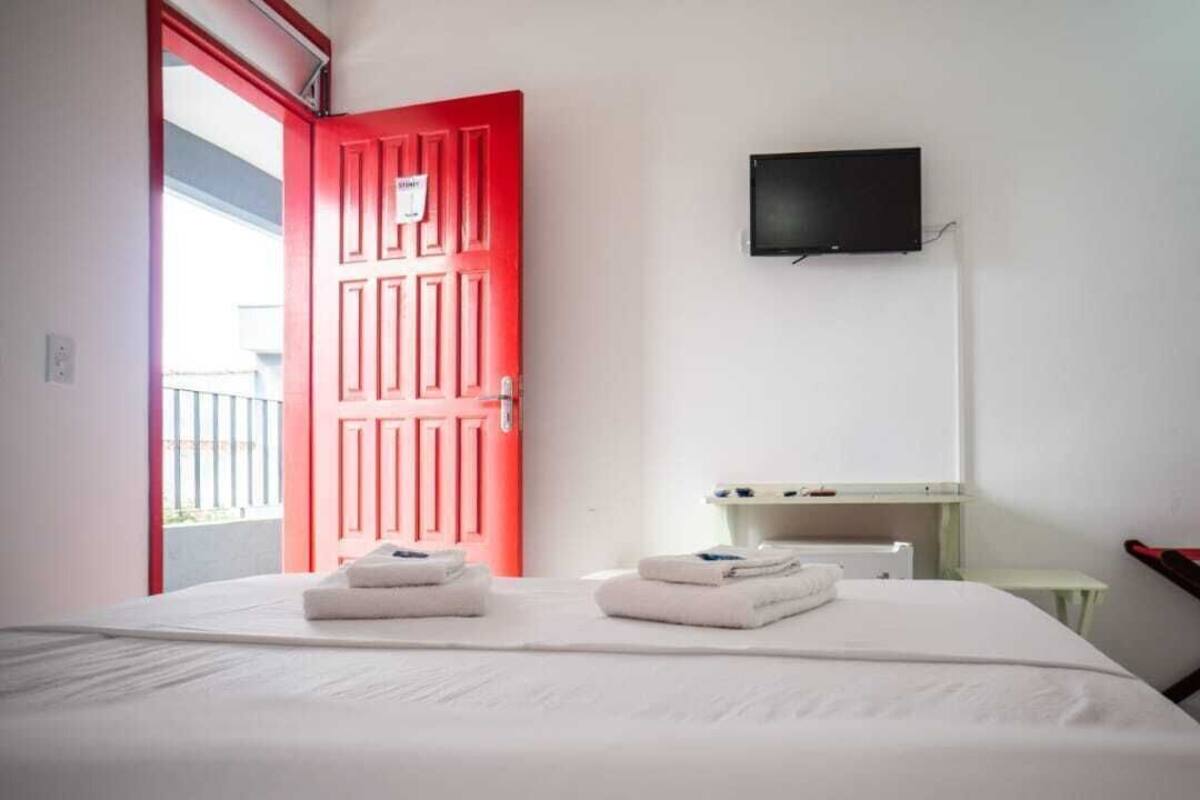 A hotel room with a red door, a flat-screen TV, a bed with two folded towels, a small table, and a chair.