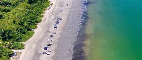 An aerial view of a beach with rows of parked cars and beach umbrellas, a clear demarcation between the sand and the water, and a line of trees in the background.