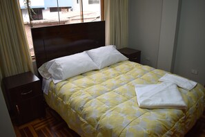 A neatly made bed with a yellow and white checkered bedspread, two white pillows, and a folded white towel on the bed. There are two bedside tables, one on each side of the bed. The room has a window with curtains, and the walls are painted a light color. - Wuasi Hospedaje - Adults only (Cusco)