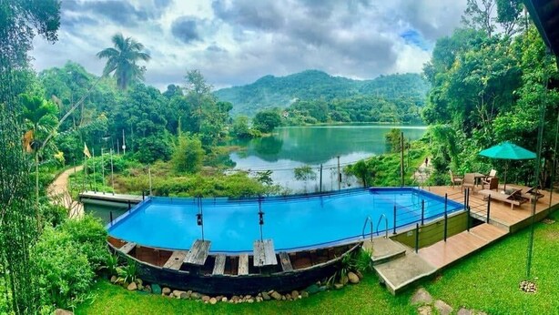 A swimming pool surrounded by lush greenery and a lake, with a deck and outdoor seating area. - Huluganga Ridge (Teldeniya)