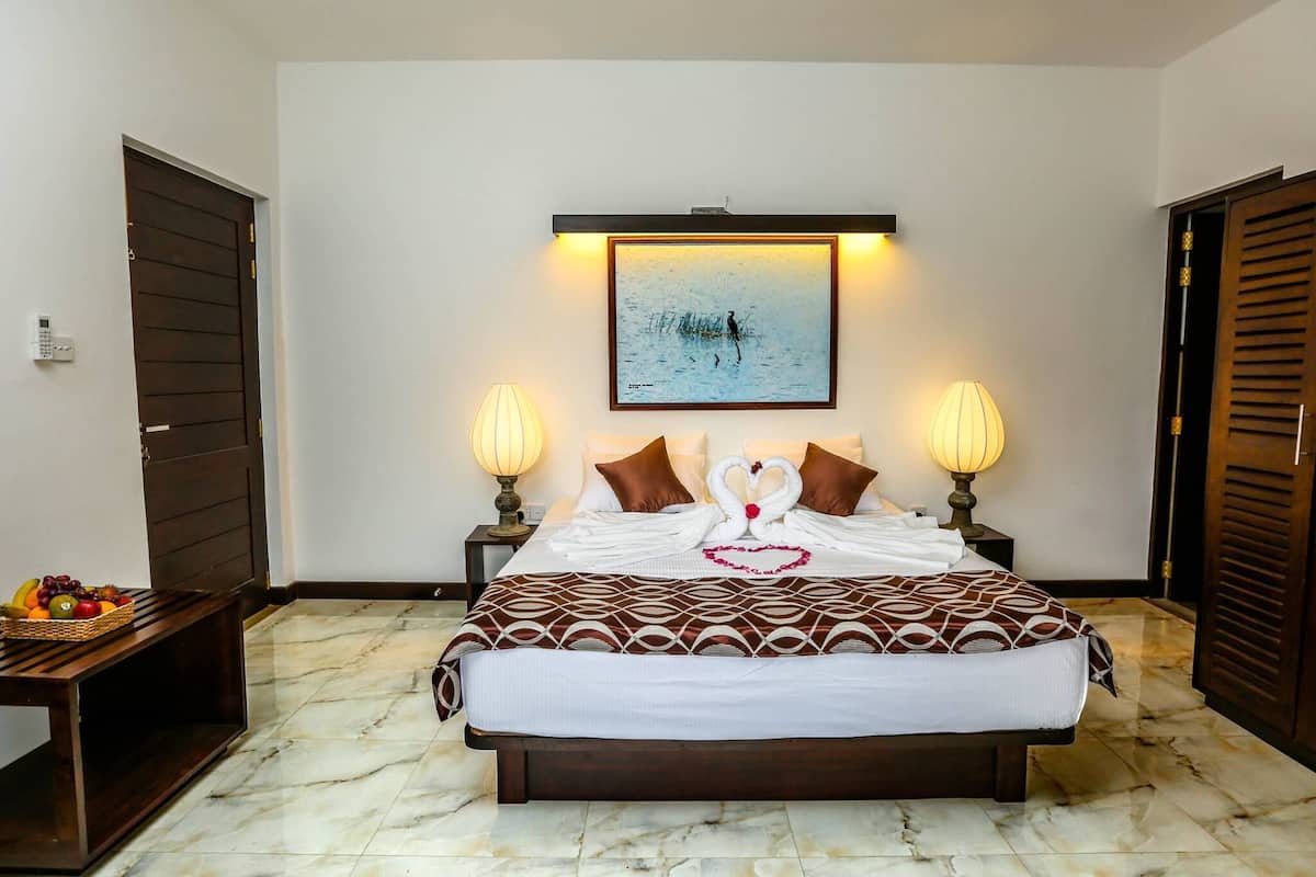 A neatly arranged hotel room with a large bed, two bedside lamps, a framed artwork above the bed, and a fruit basket on a small table beside the bed.