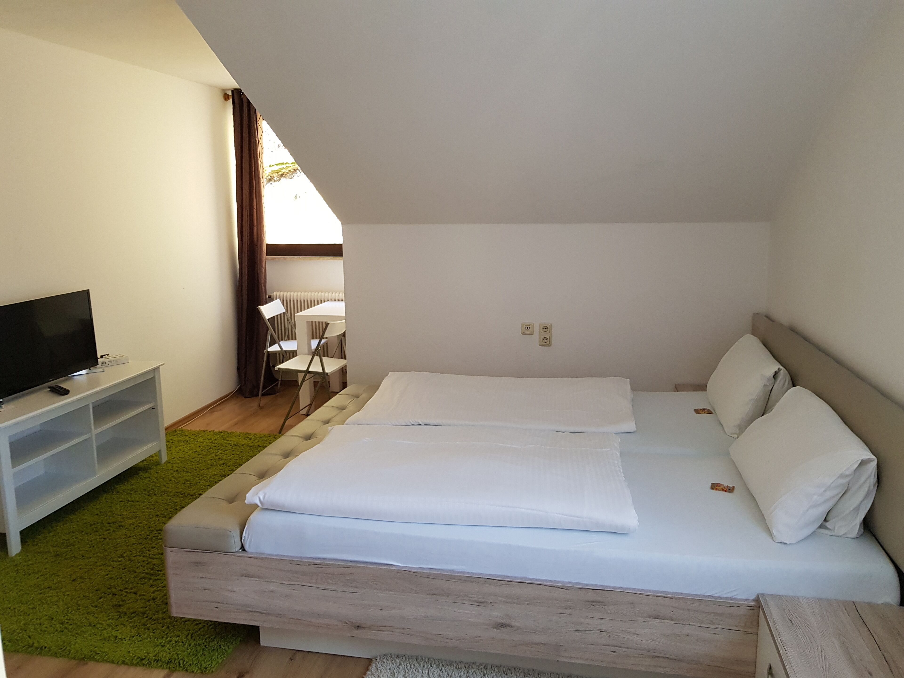 A bedroom with a bed, a TV on a stand, a small table, and a chair.