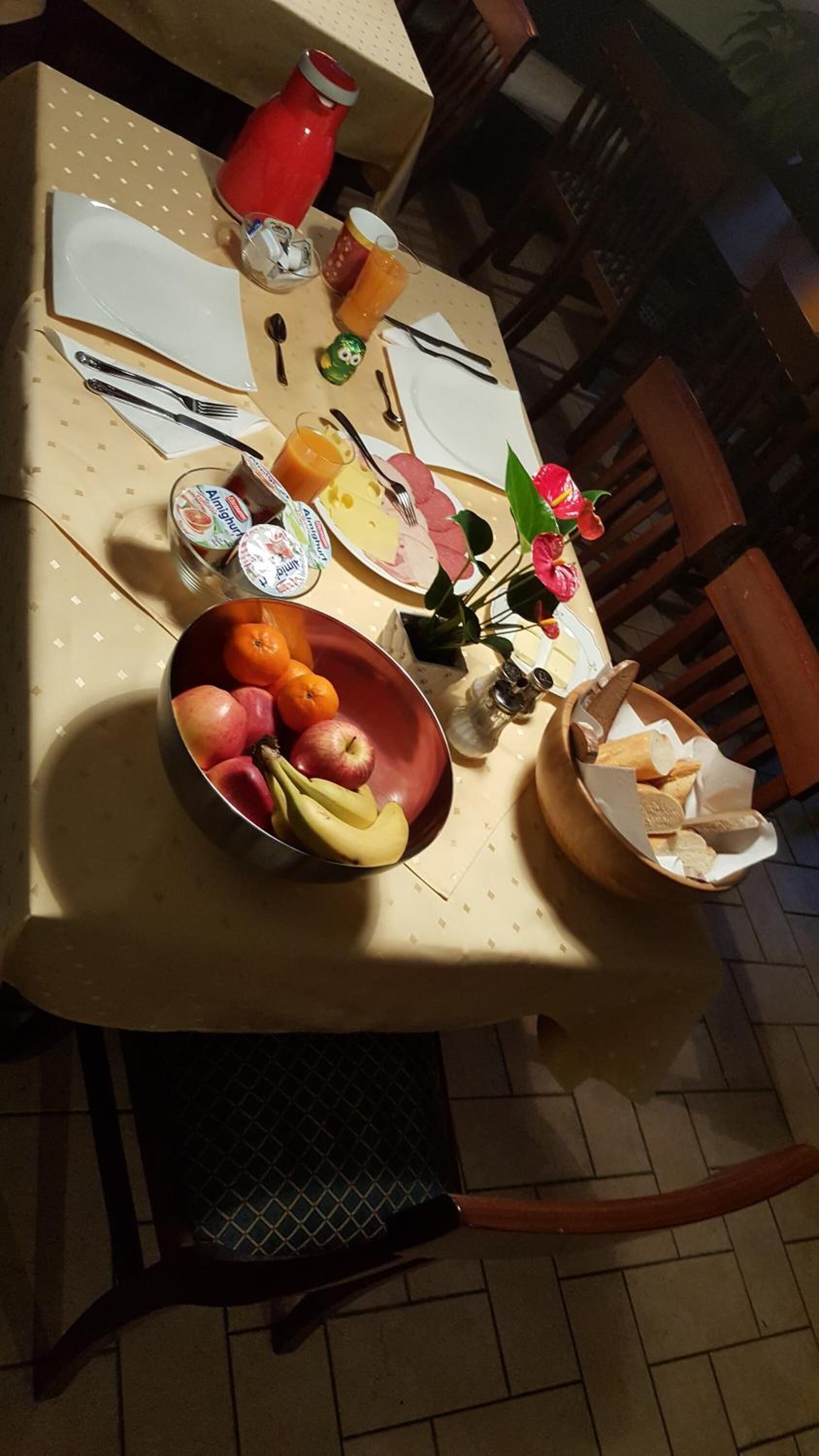 Una mesa de comedor puesta con platos, cubiertos, un cuenco de fruta, un vaso de jugo y un florero con flores.