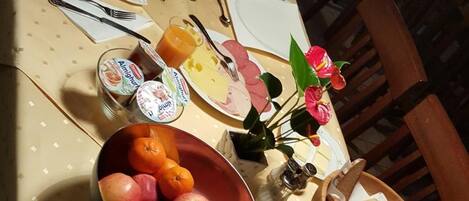 Una mesa de comedor puesta con platos, cubiertos, un cuenco de fruta, un vaso de jugo y un florero con flores.