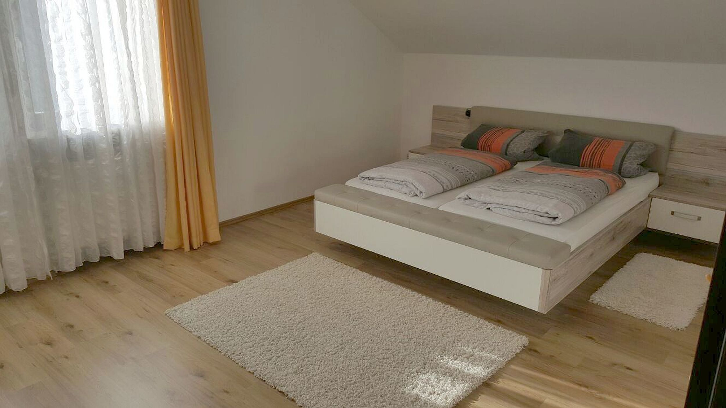 Ein Schlafzimmer mit einem Bett, einem Nachttisch, einem Fenster mit Vorhängen und Holzboden.