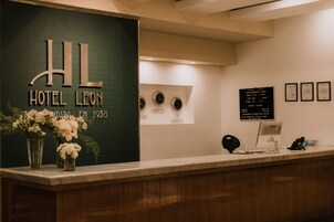 Een hotel receptie met een bordje met de tekst 'Hotel Leon' en een jaartal '1938'. Er hangen klokken aan de muur en staat een computer op het bureau.