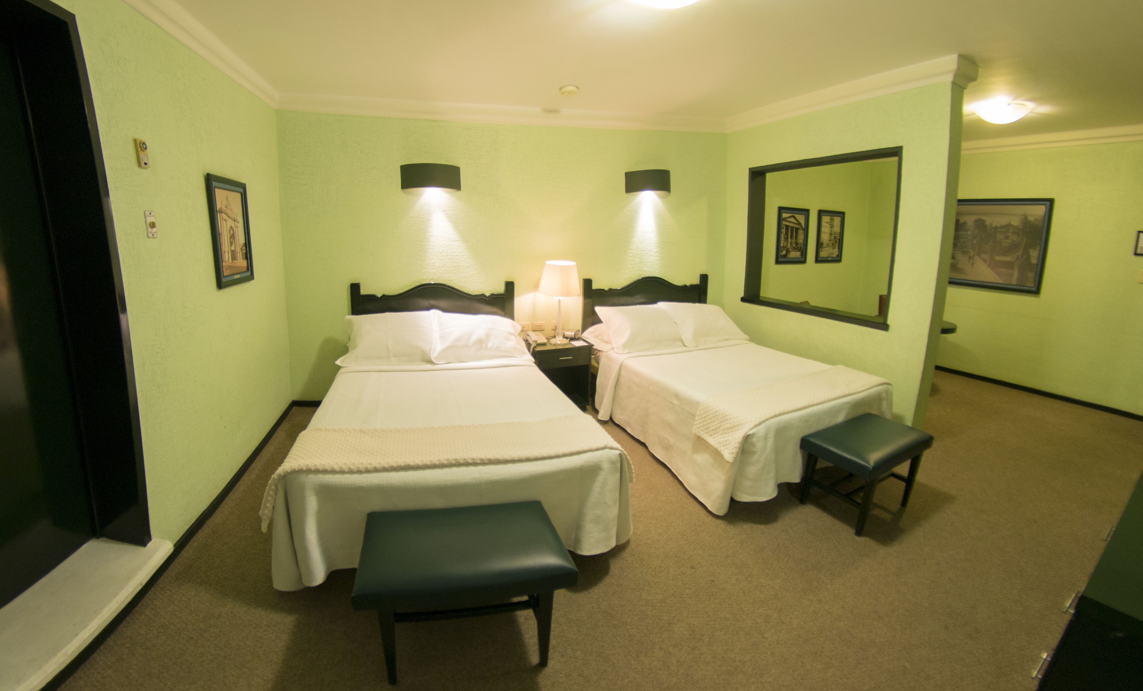 A hotel room with two single beds, a bench, a nightstand with a lamp, and framed pictures on the walls.