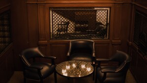 A room with a round table and three chairs, a window with a decorative grille, and wooden paneling on the walls. - Hotel Leon (León)