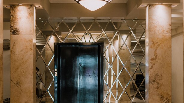 A lobby with a central black door, marble columns, a geometric patterned wall, and a circular reflective floor. - Hotel Leon (León)