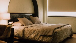 A neatly made bed with patterned bedding, a bedside lamp, and a chair in a room with a curved headboard and a window. - Hotel Leon (León)