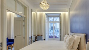 A large bed with white linens, a chandelier, and a view of a balcony with railings.