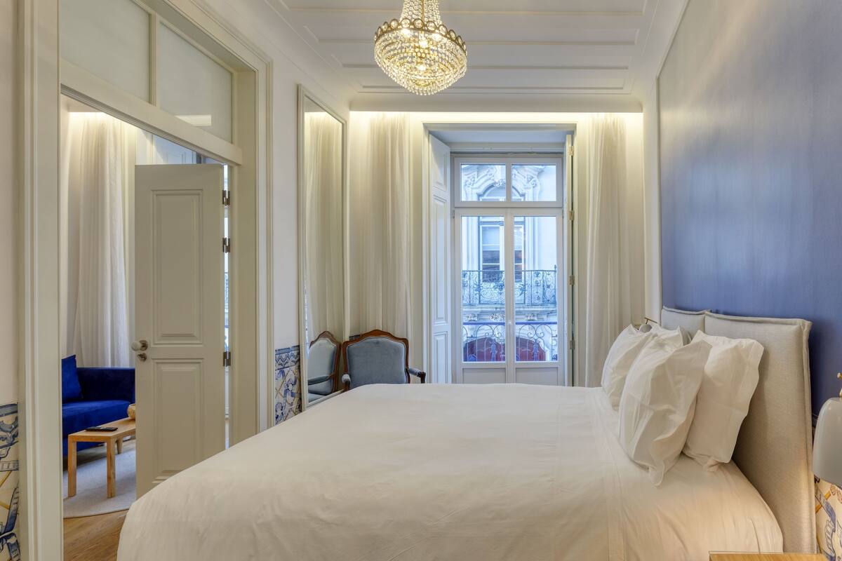 Un grand lit avec du linge de lit blanc, un lustre et une vue sur un balcon doté de balustrades.
