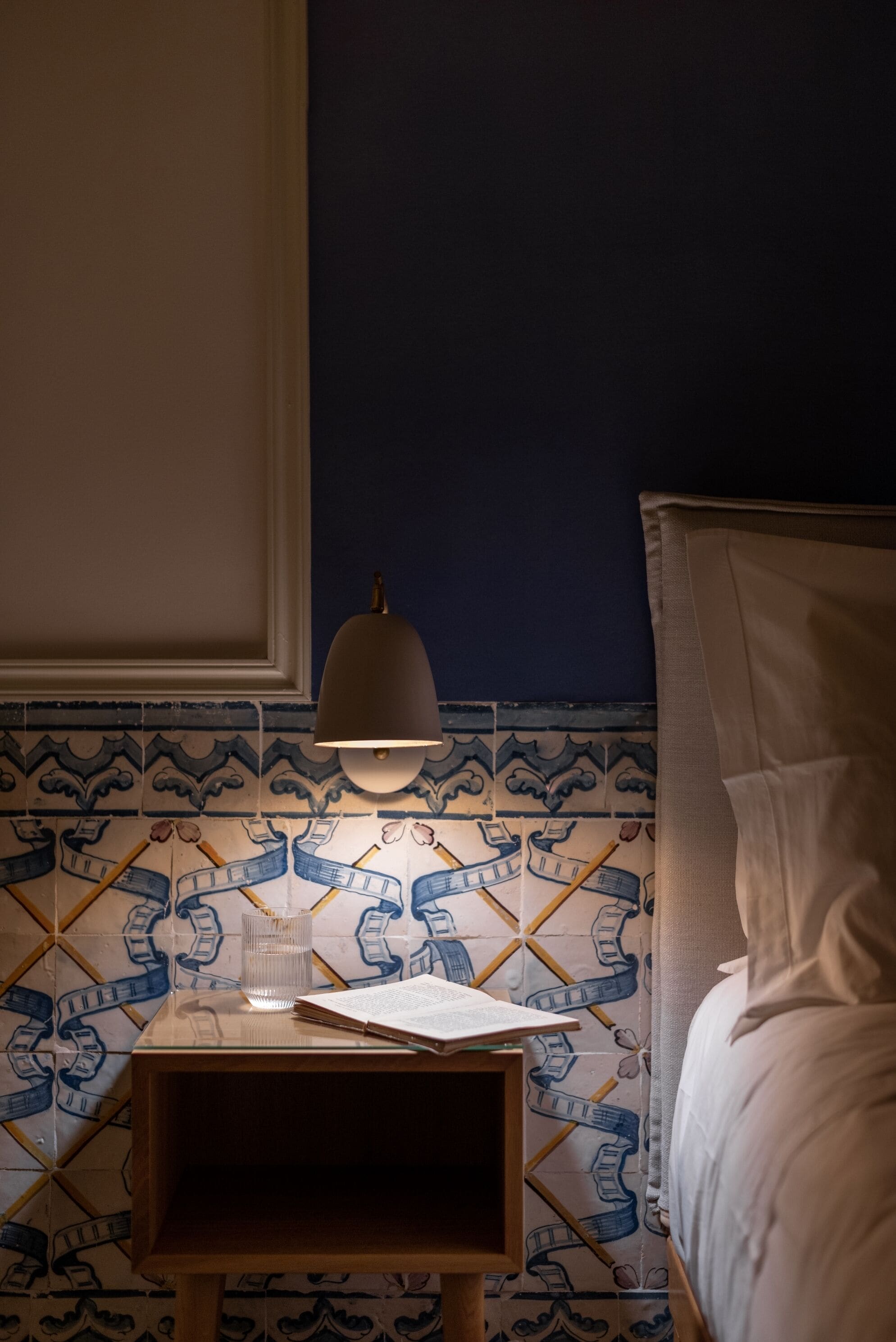 Una cama bien hecha con cabecera, una mesita de noche con lámpara, y una pared de azulejos con un diseño de historieta.