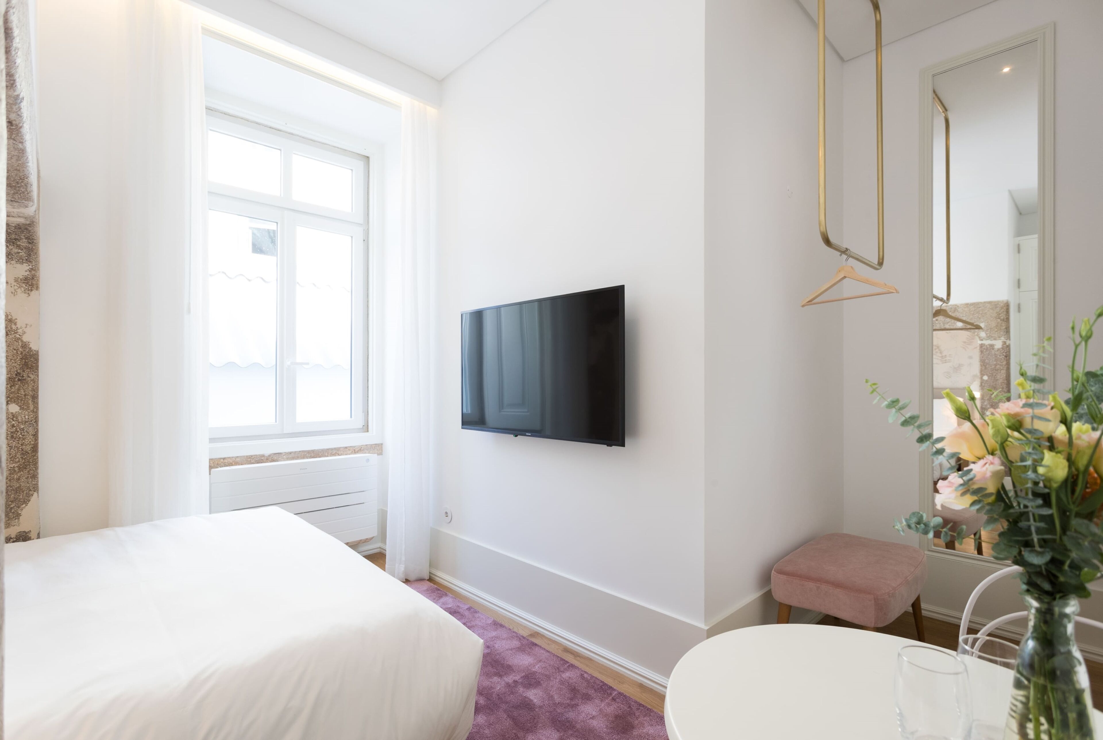 Une chambre d’hôtel avec un lit, un téléviseur fixé au mur, une petite table avec un vase de fleurs et une fenêtre avec des rideaux.