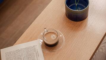 Uma mesa de madeira com uma chávena de vidro com café, um livro e um vaso azul com plantas secas.